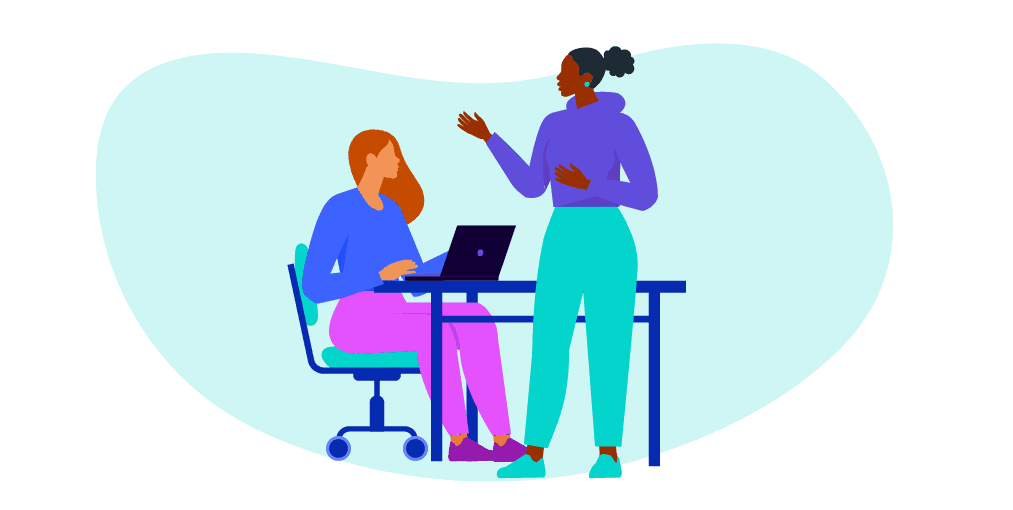 Two female colleagues are discussing work. One is at the work dest and the other is standing opposite.